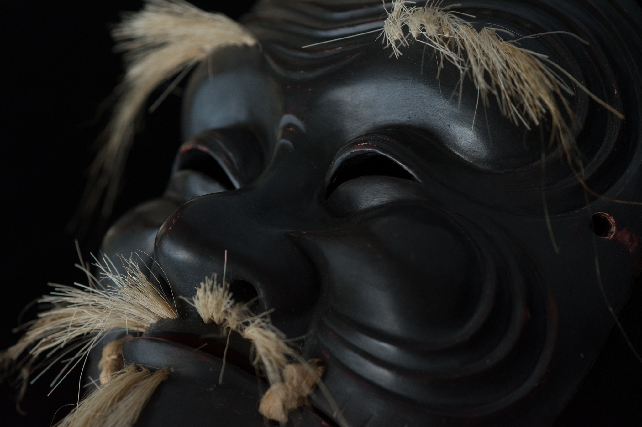 Three-quarter view of a black Kokushiki-jō Noh mask with pale hair and cord attachments